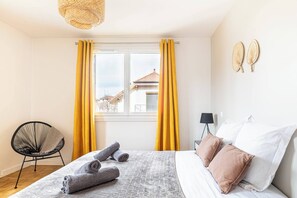 Um quarto com cama, cadeira, janela com cortinas e vista para prédios.