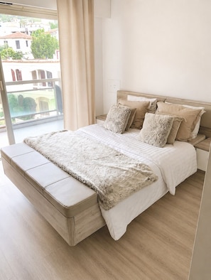Une chambre à coucher moderne avec un grand lit, une tête de lit en bois et une vue sur les immeubles par la fenêtre.