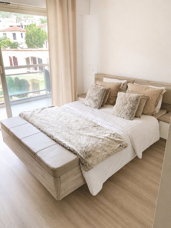A modern bedroom with a large bed, a wooden headboard, and a view of buildings through a window.