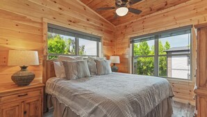 A wooden cabin bedroom with a bed, two nightstands, a ceiling fan, and large windows offering a view of greenery outside. - Amazing 4BD Lodge w/ Hot Tub! Games + NEAR TOWN! (Pigeon Forge)