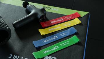 Élastiques de résistance de la marque MDBuddy, disponibles avec différents niveaux de résistance (léger, moyen, fort, très fort), ainsi qu’un pistolet de massage sur un tapis.