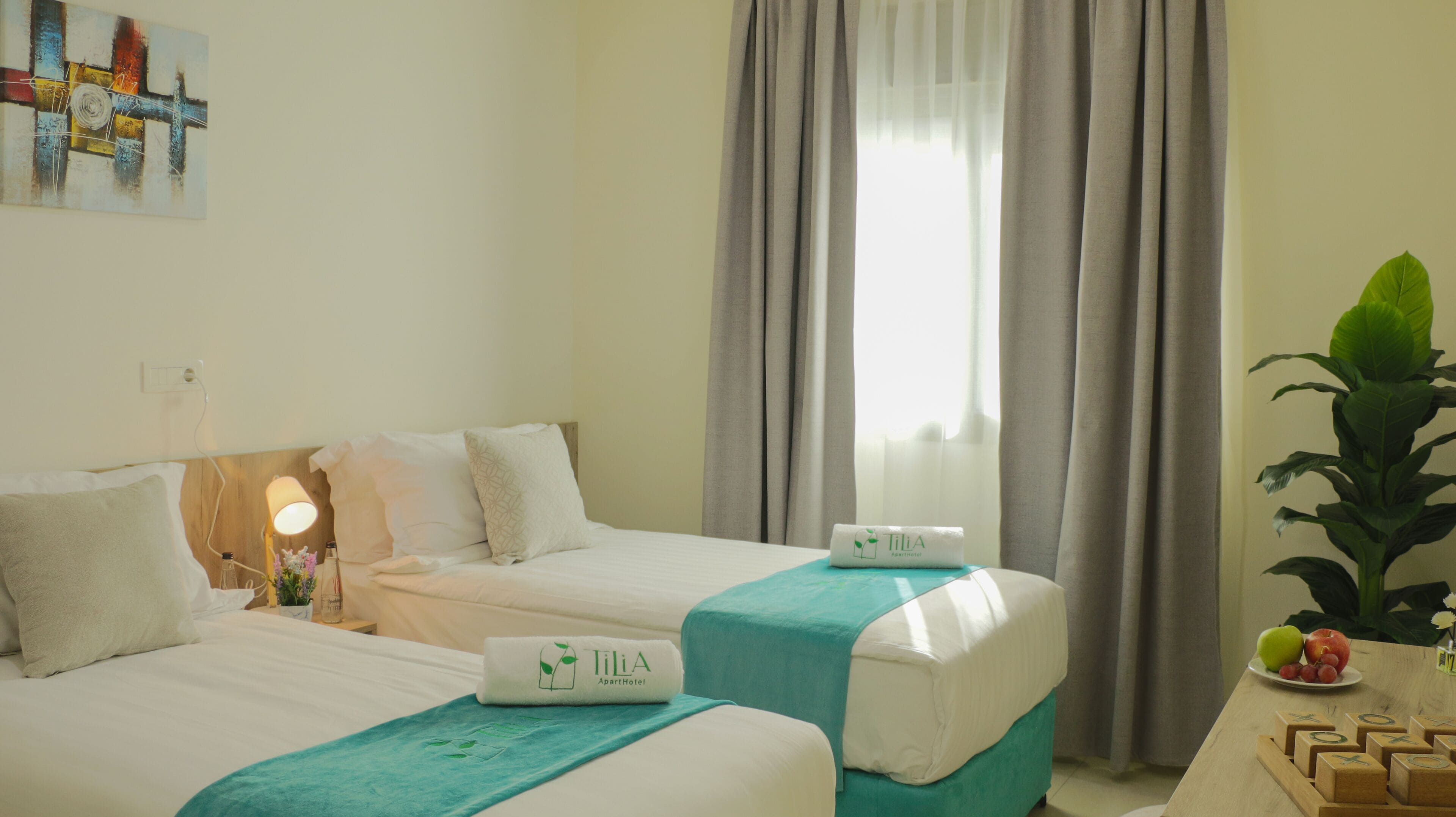 Une chambre d’hôtel avec deux lits, une fenêtre avec des rideaux, une plante et une table avec des fruits.