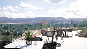 Una terraza en la azotea con mobiliario exterior, barandilla de cristal y vistas a la ciudad.