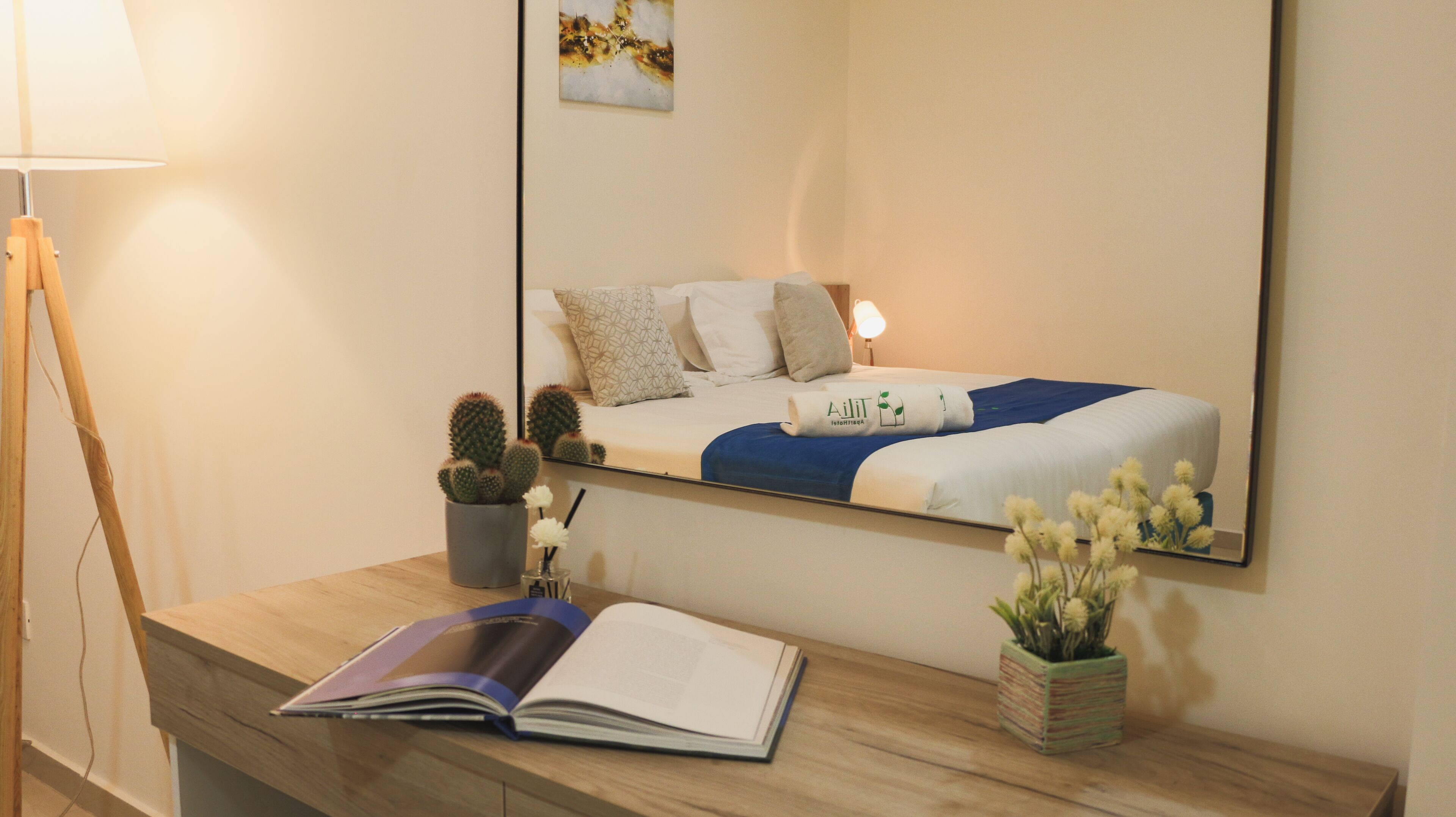 A hotel room with a bed, a bedside table with a book, a lamp, and a potted plant.