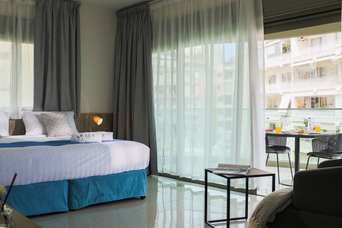 Quarto de hotel moderno com uma cama grande, uma mesa pequena com uma cadeira e uma varanda com área de refeições.