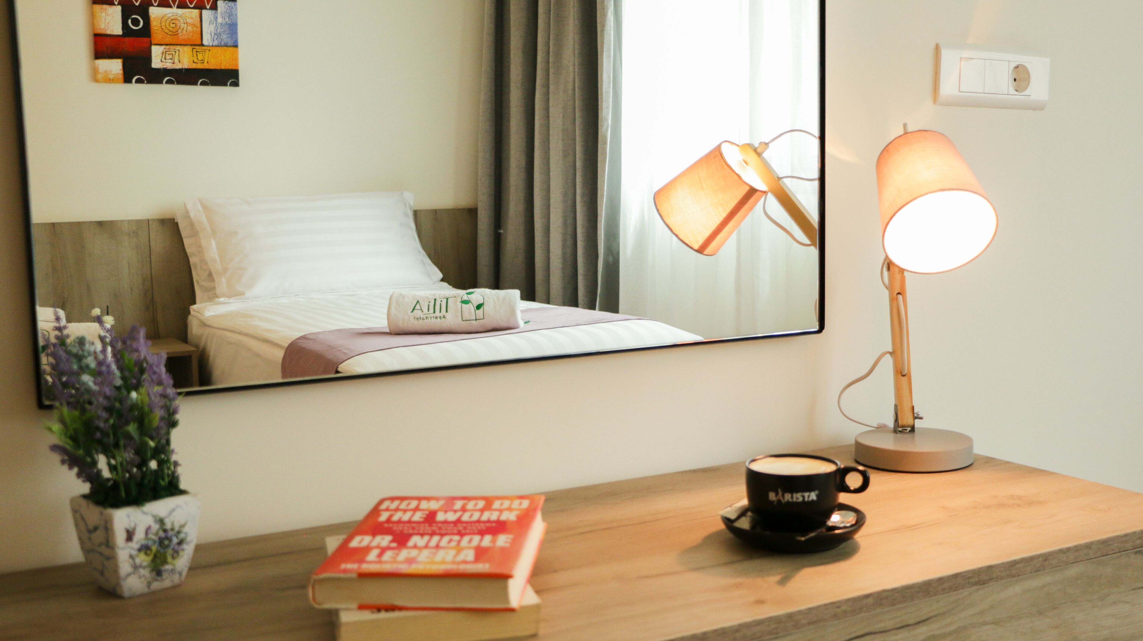 A hotel room with a bed, a bedside lamp, a coffee cup, a book, and a mirror reflecting the room.