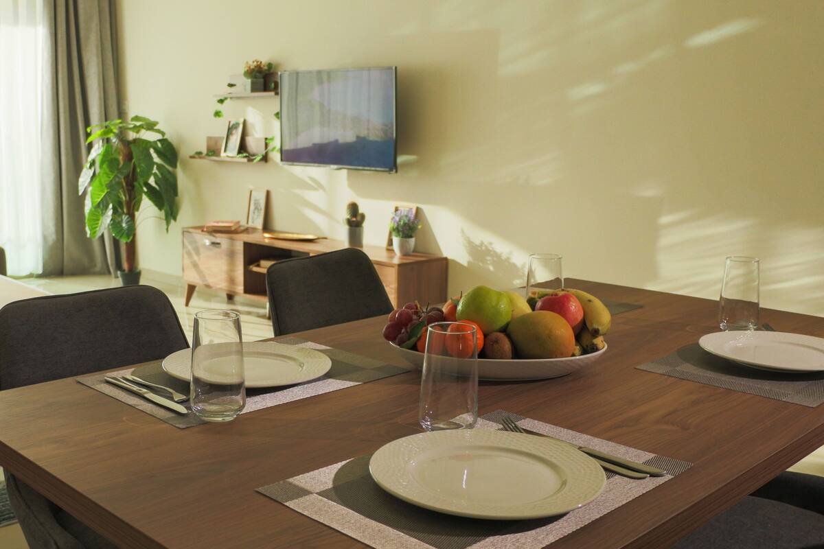 Uma área de refeições com uma mesa de madeira posta para quatro pessoas, incluindo pratos, talheres e uma tigela de frutas.