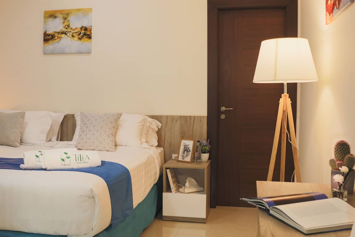 Quarto de hotel com cama, almofadas, mesa de cabeceira com abajur, porta e um quadro na parede.