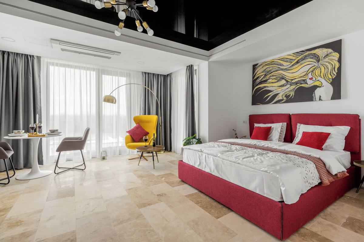 Ein modernes Hotelzimmer mit einem roten Bett, einem Essbereich mit Tisch und Stühlen sowie einem großen Fenster mit Vorhängen.