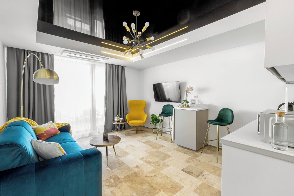 Ein modernes Wohnzimmer mit einem blauen Sofa, einem gelben Sessel und einem grünen Stuhl. Ein kleiner Tisch mit Lampe, Fernseher und einer Küchenzeile mit Spüle und Arbeitsplatte.