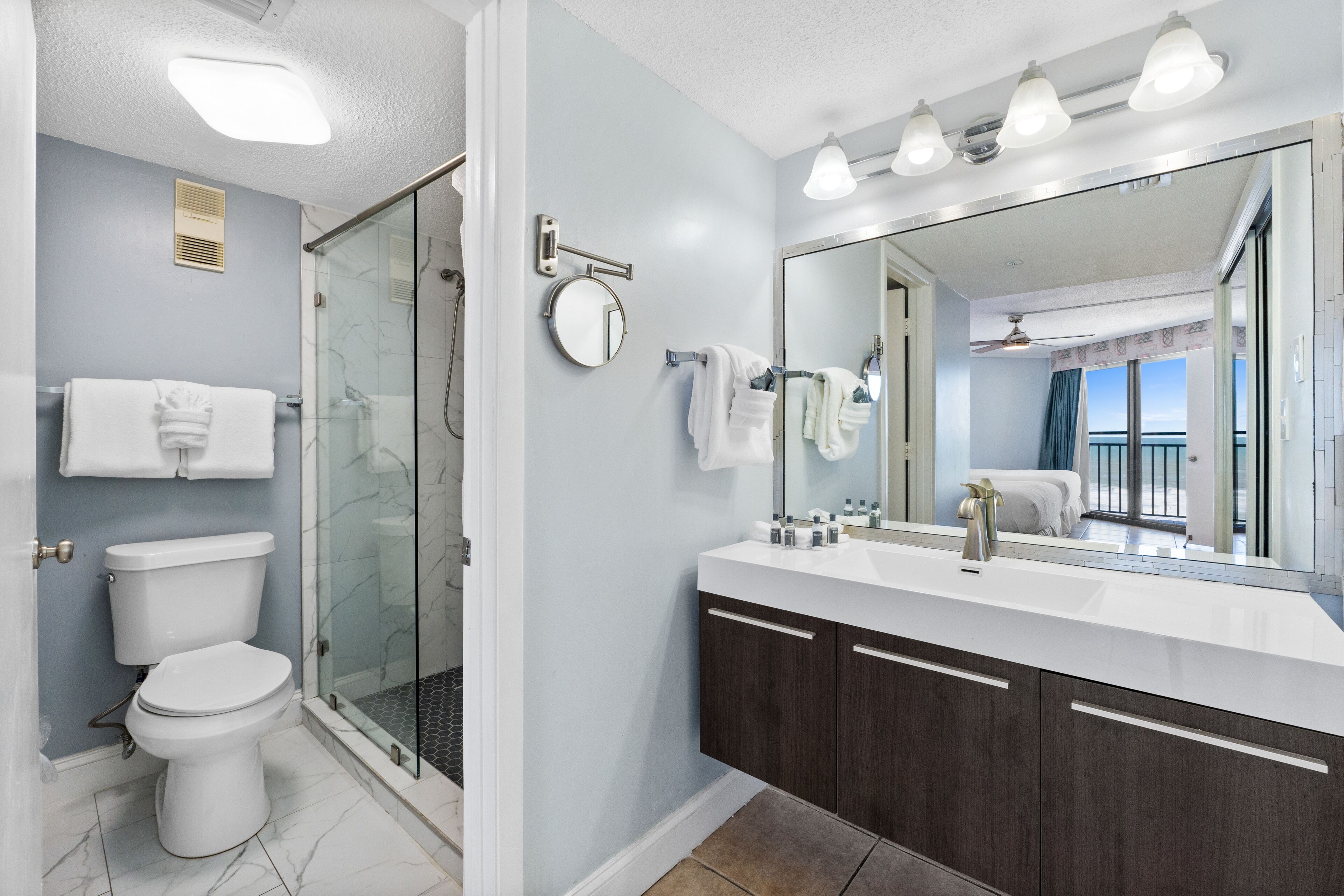Un baño moderno con ducha envuelta en vidrio, inodoro blanco, espejo grande y tocador con armarios de madera oscura.