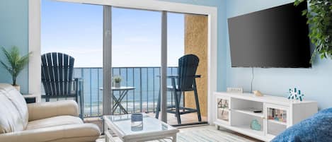 A living room with a large TV, a sofa, a coffee table, and a view of the ocean through a sliding glass door.