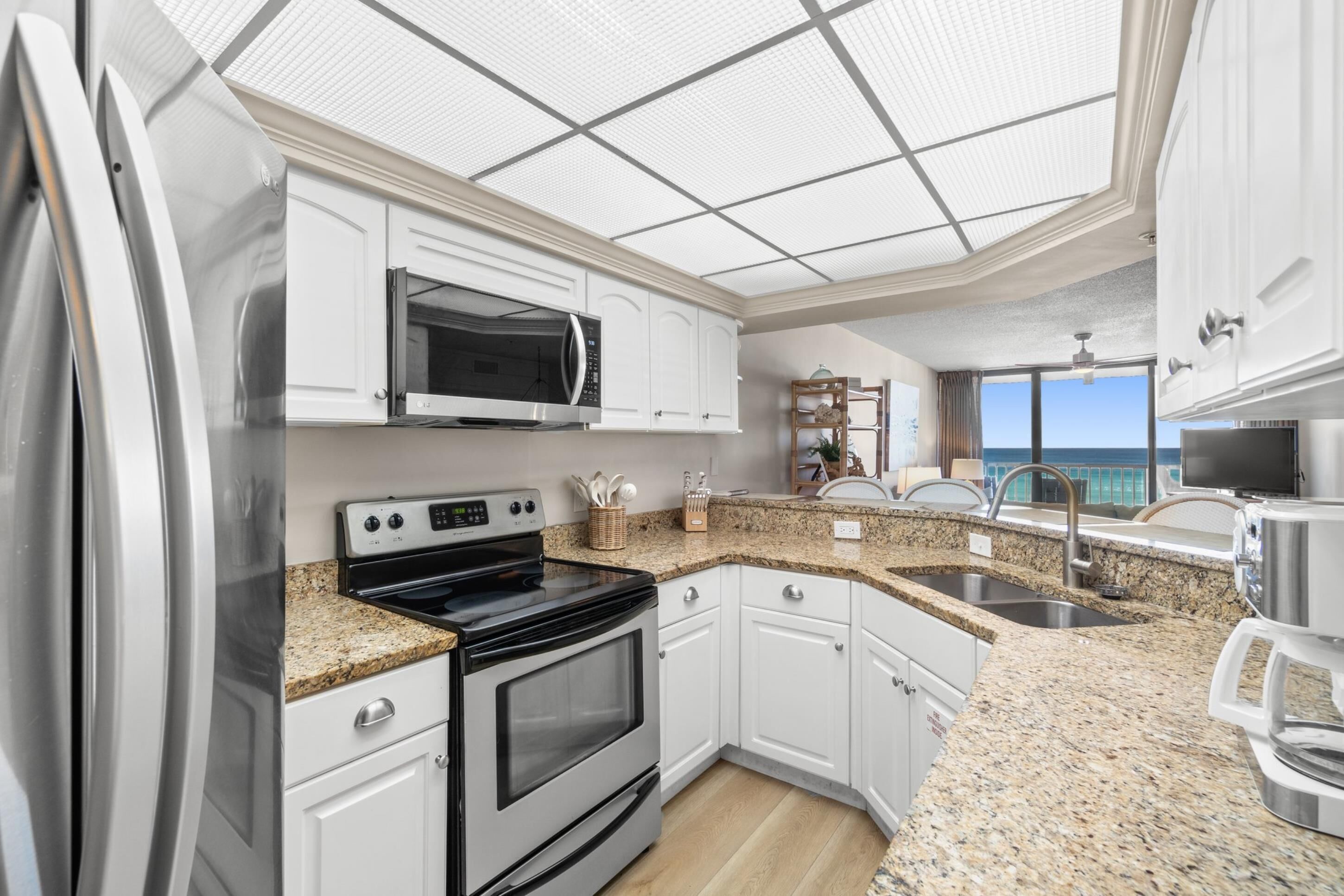 A modern kitchen with white cabinets, granite countertops, stainless steel appliances, and a large window overlooking the ocean.