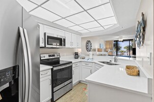 Une cuisine moderne avec des meubles blancs, des appareils en acier inoxydable, un grand îlot central doté d’un évier et une vue sur l’extérieur grâce à une porte-fenêtre coulissante.