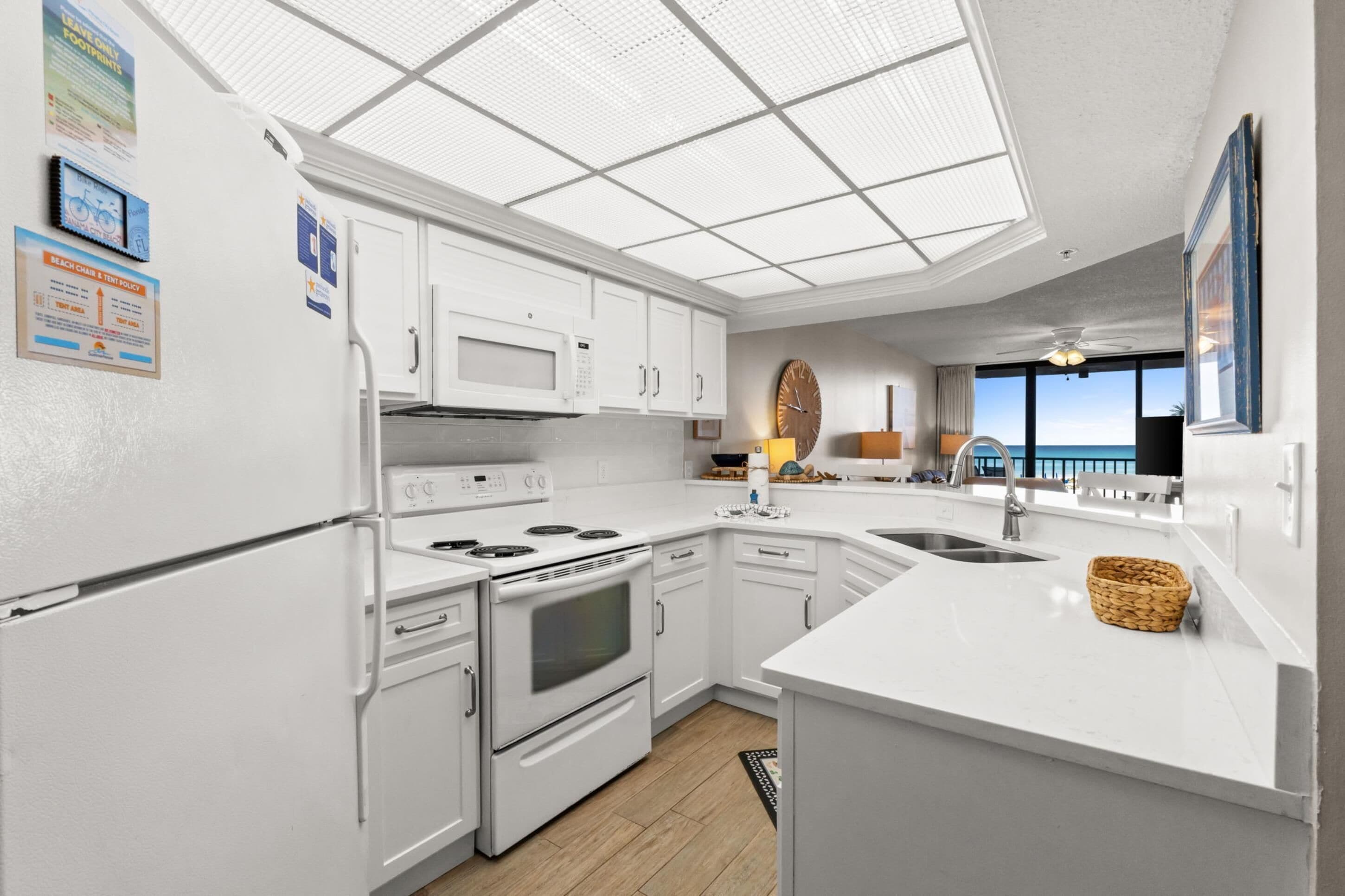 Eine moderne Küche mit weißen Schränken, integriertem Mikrowellenherd, Herd und Backofen, einer großen Kochinsel mit Spüle und Blick aufs Meer durch große Fenster.