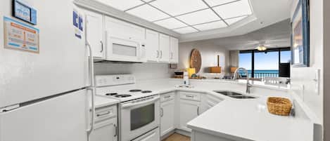 A modern kitchen with white cabinetry, a built-in microwave, stove, and oven, a large island with a sink, and a view of the ocean through large windows.