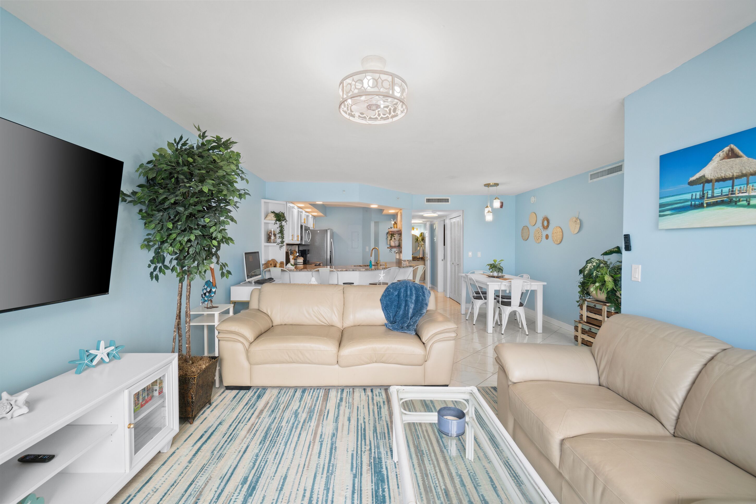 Ein modernes Wohnzimmer mit einem beigen Sofa, einem Fernseher, einer Chandelieber und einem Essbereich im Hintergrund.
