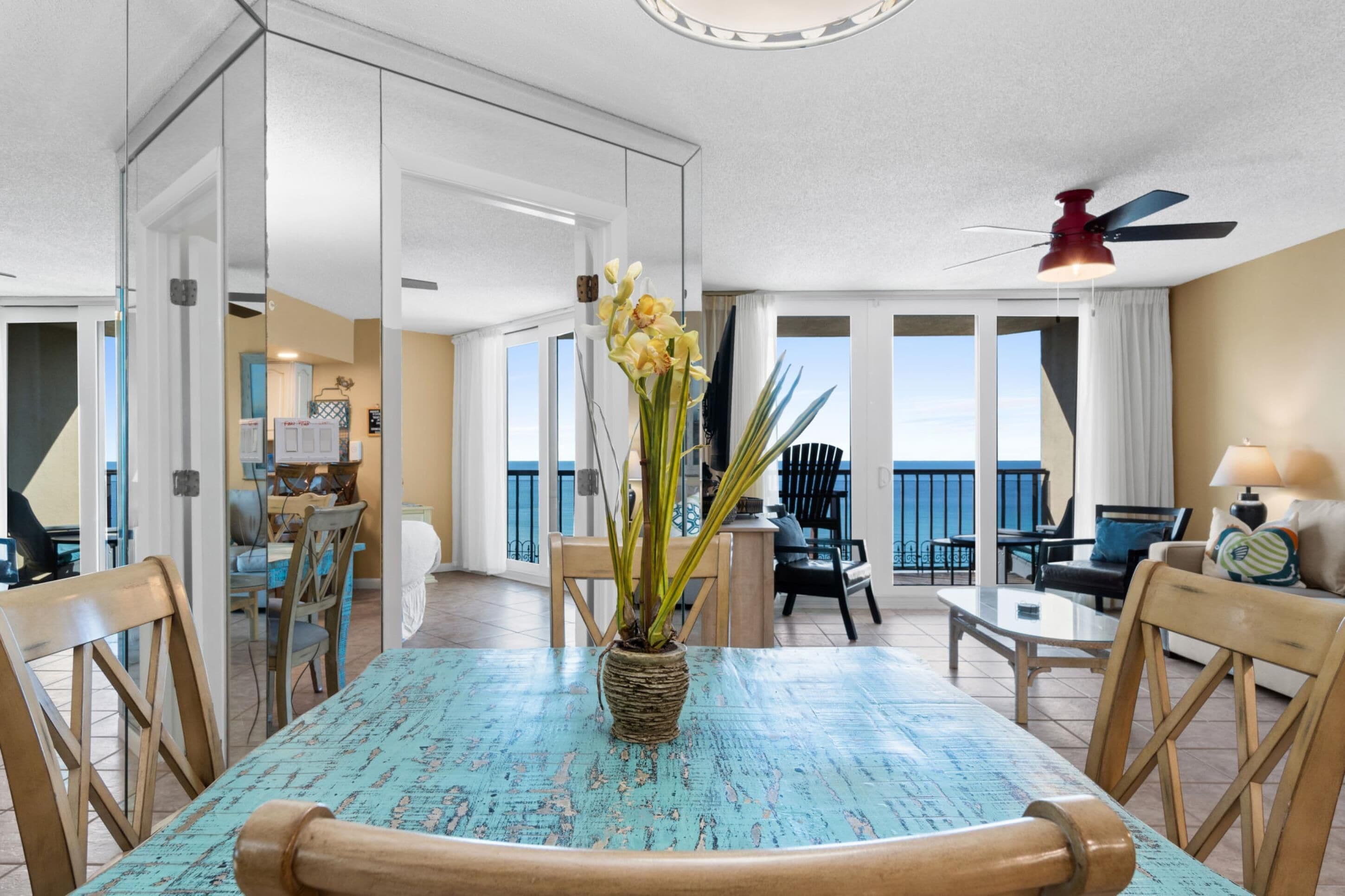 A dining area with a table, chairs, and a vase of flowers. There is a balcony with a view of the sky.