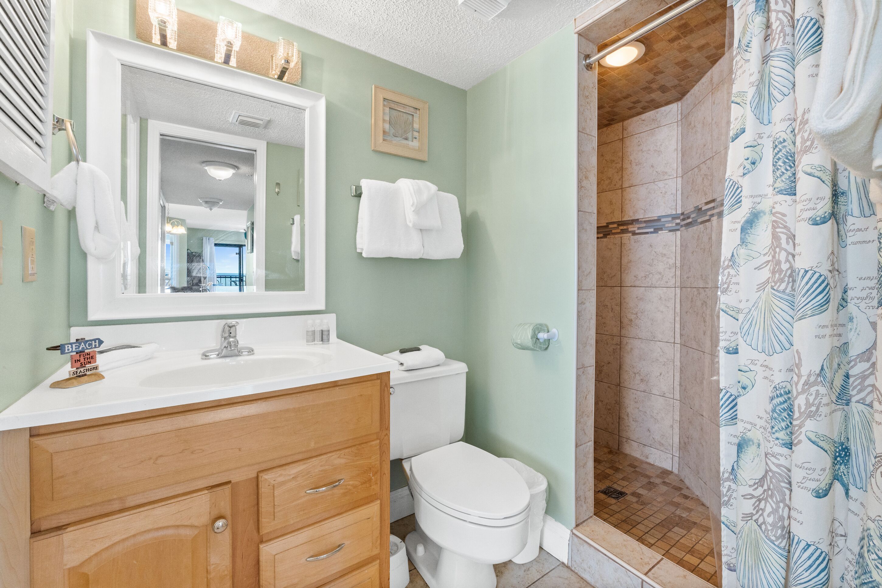 A bathroom with a large mirror, white sink, wooden vanity, white toilet, and a shower with a leaf-patterned curtain.