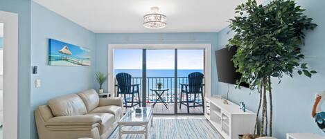 A living room with a sofa, a coffee table, a TV, and a balcony with chairs and a plant.