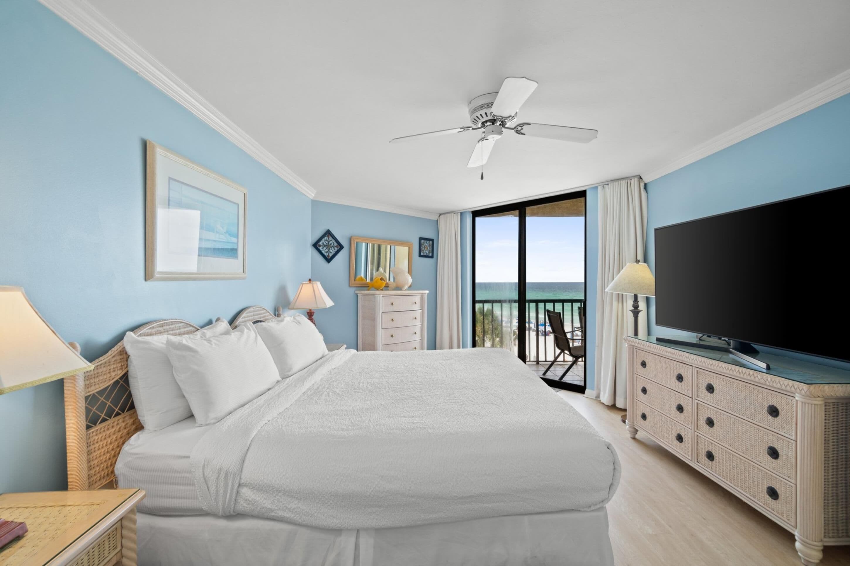 A hotel room with a large bed, a television, a ceiling fan, a balcony with a view, and a dresser.
