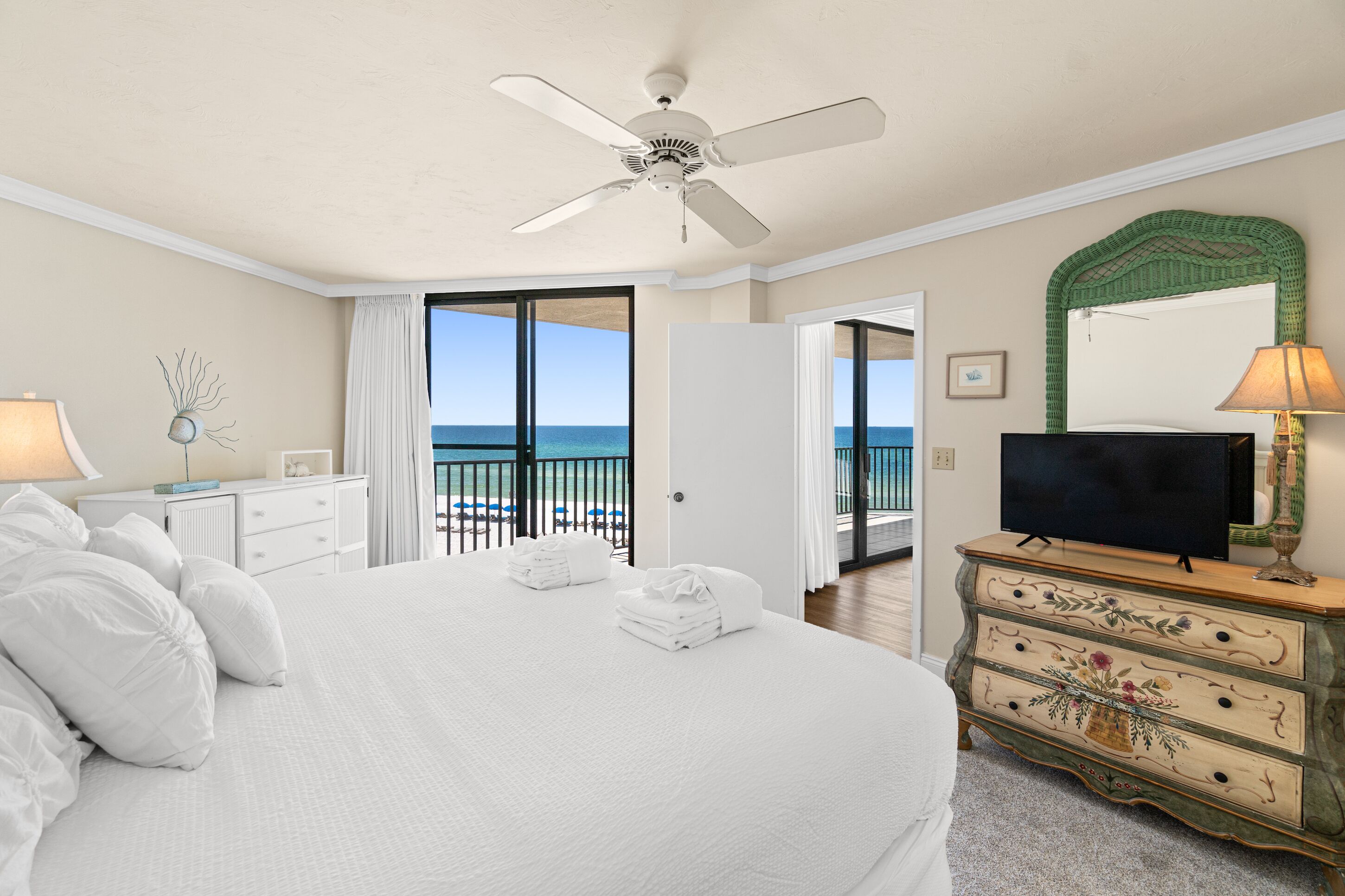 A hotel room with a large bed, a television, a dresser, and a balcony with a view of the ocean.