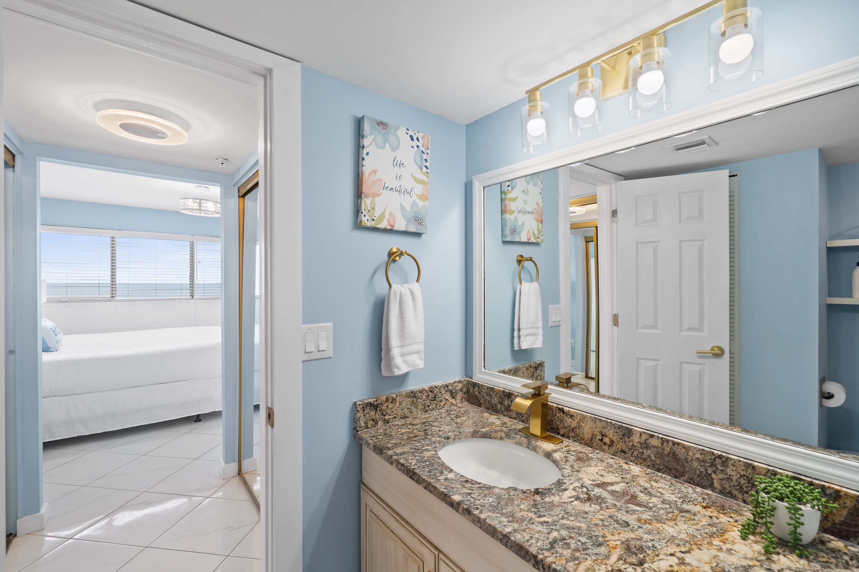 Une salle de bain avec un lavabo blanc, un comptoir en granit et un grand miroir. On y trouve un porte-serviettes avec une serviette blanche, ainsi qu’un élément décoratif mural au-dessus du miroir.