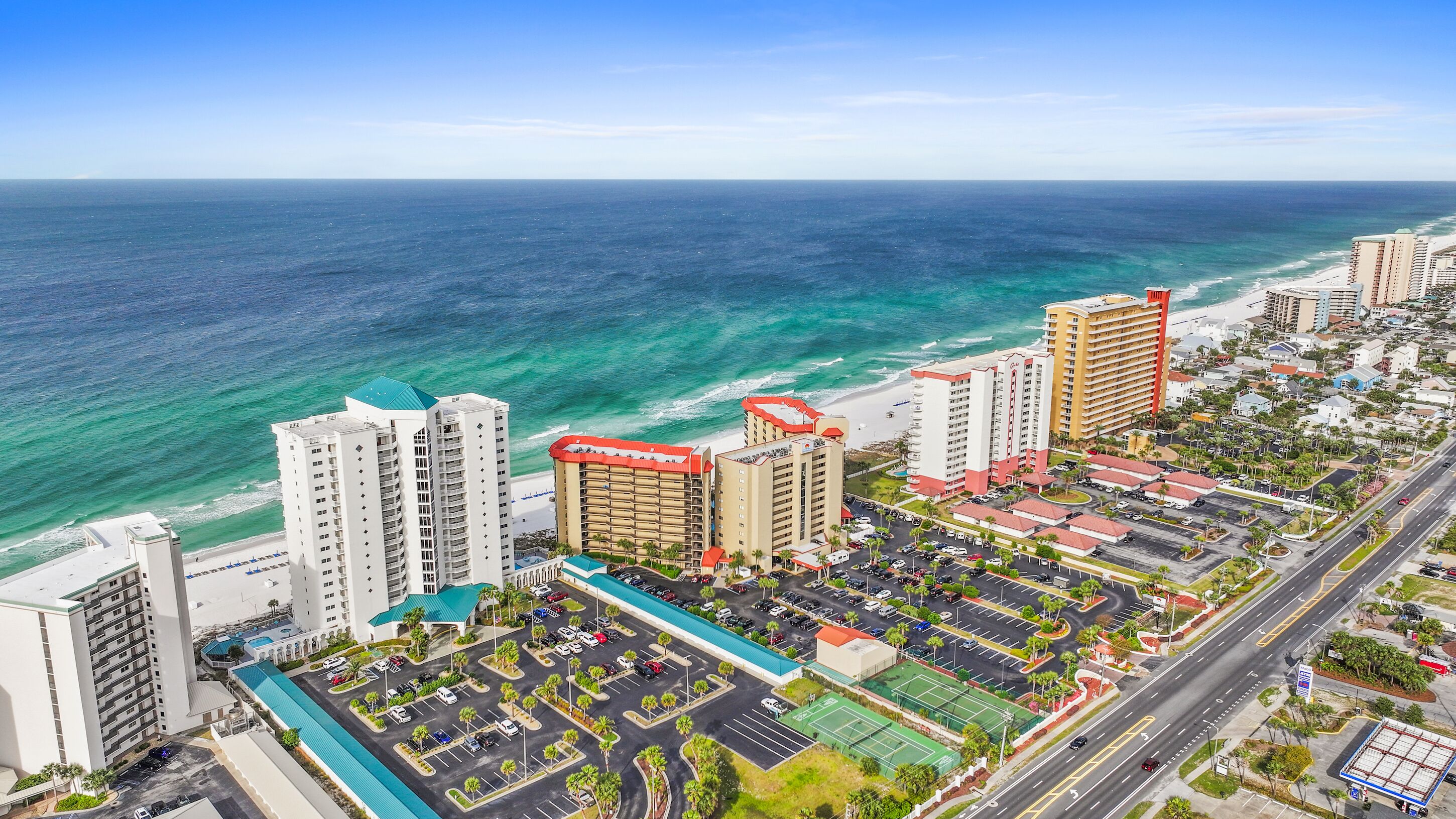 A coastal hotel with multiple high-rise buildings, a parking lot, and a beach in the background.