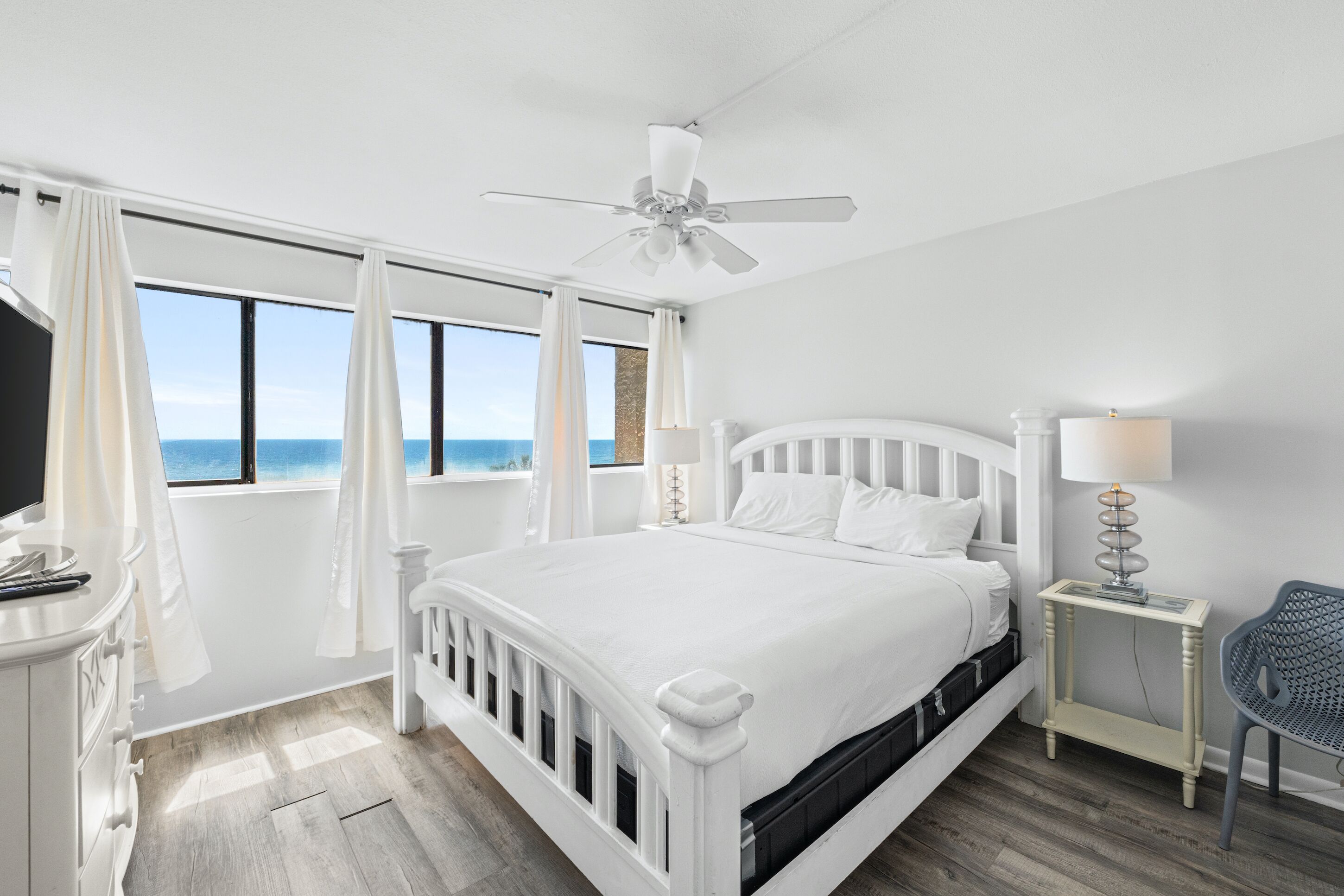 A white bedroom with a bed, a nightstand, a chair, a ceiling fan, and a view of the ocean.