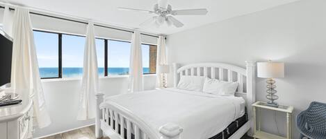 A white bedroom with a bed, a nightstand, a chair, a ceiling fan, and a view of the ocean.