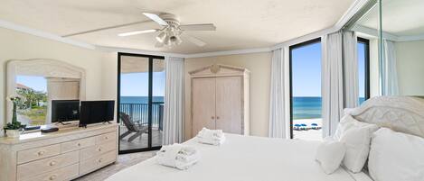 A hotel room with a large bed, a TV, a dresser, and a balcony with a view of the beach.