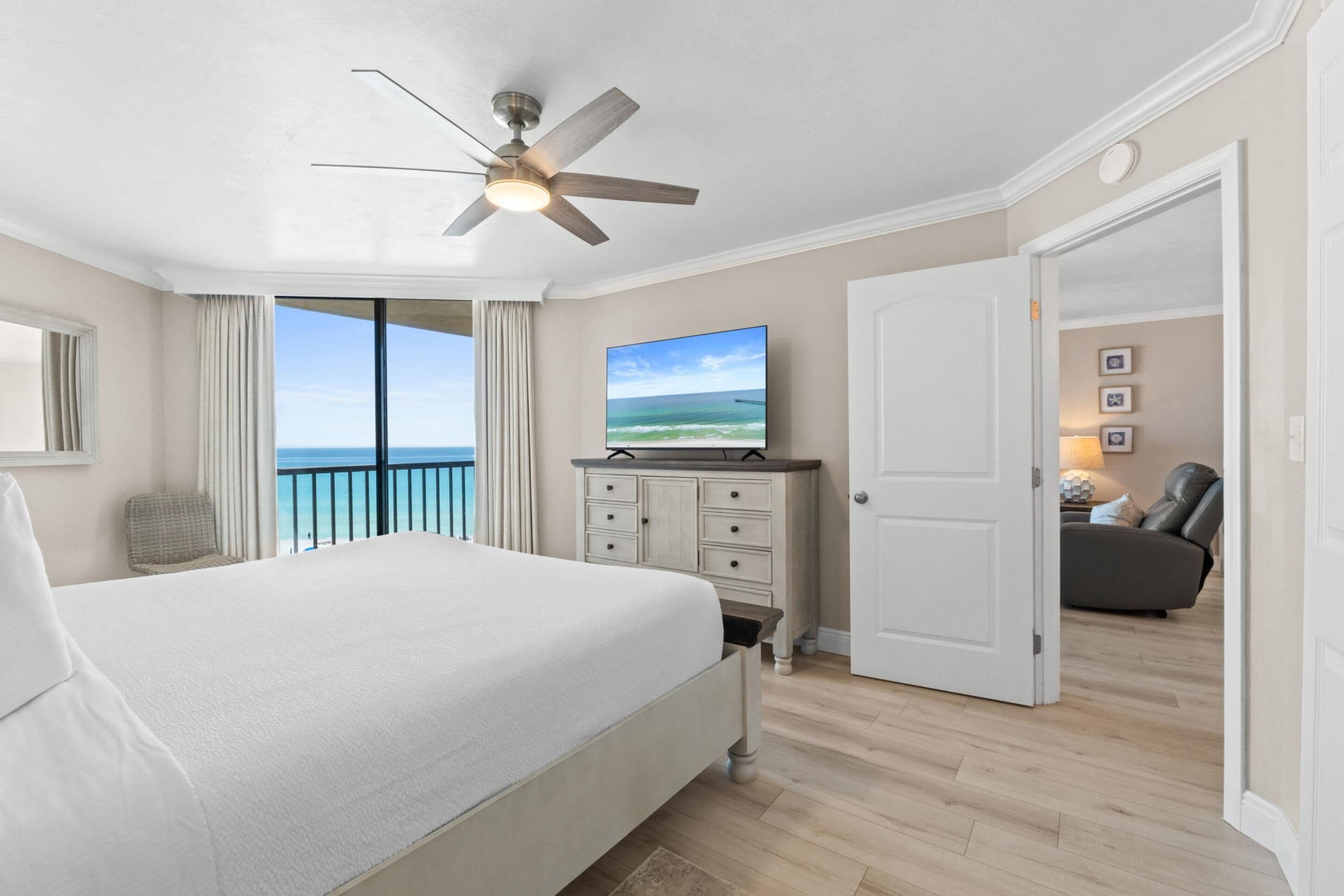 A bedroom with a bed, a ceiling fan, a TV, a dresser, and a door leading to another room.
