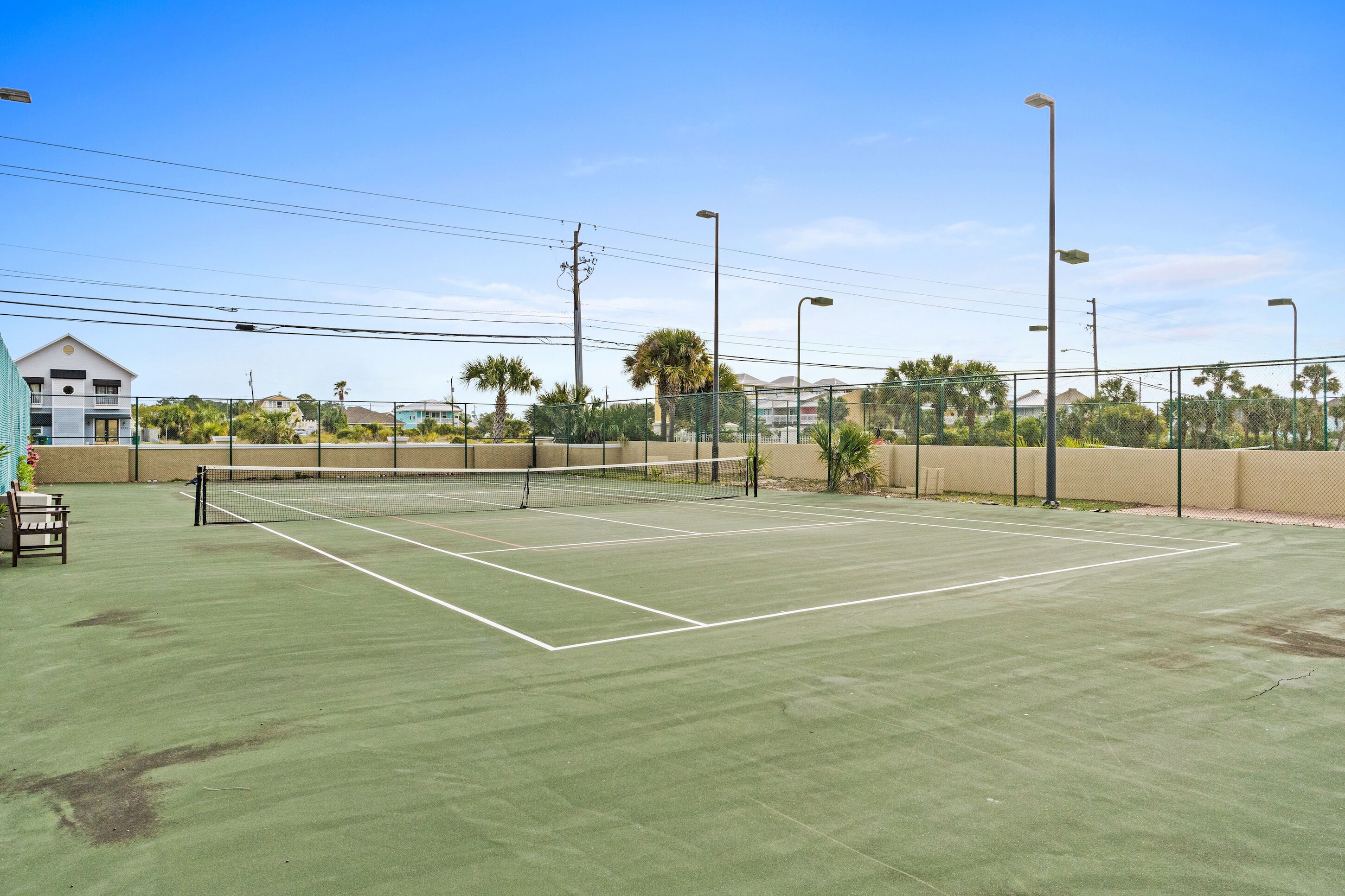 An outdoor tennis court with a net, surrounded by a fence, and palm trees in the background.