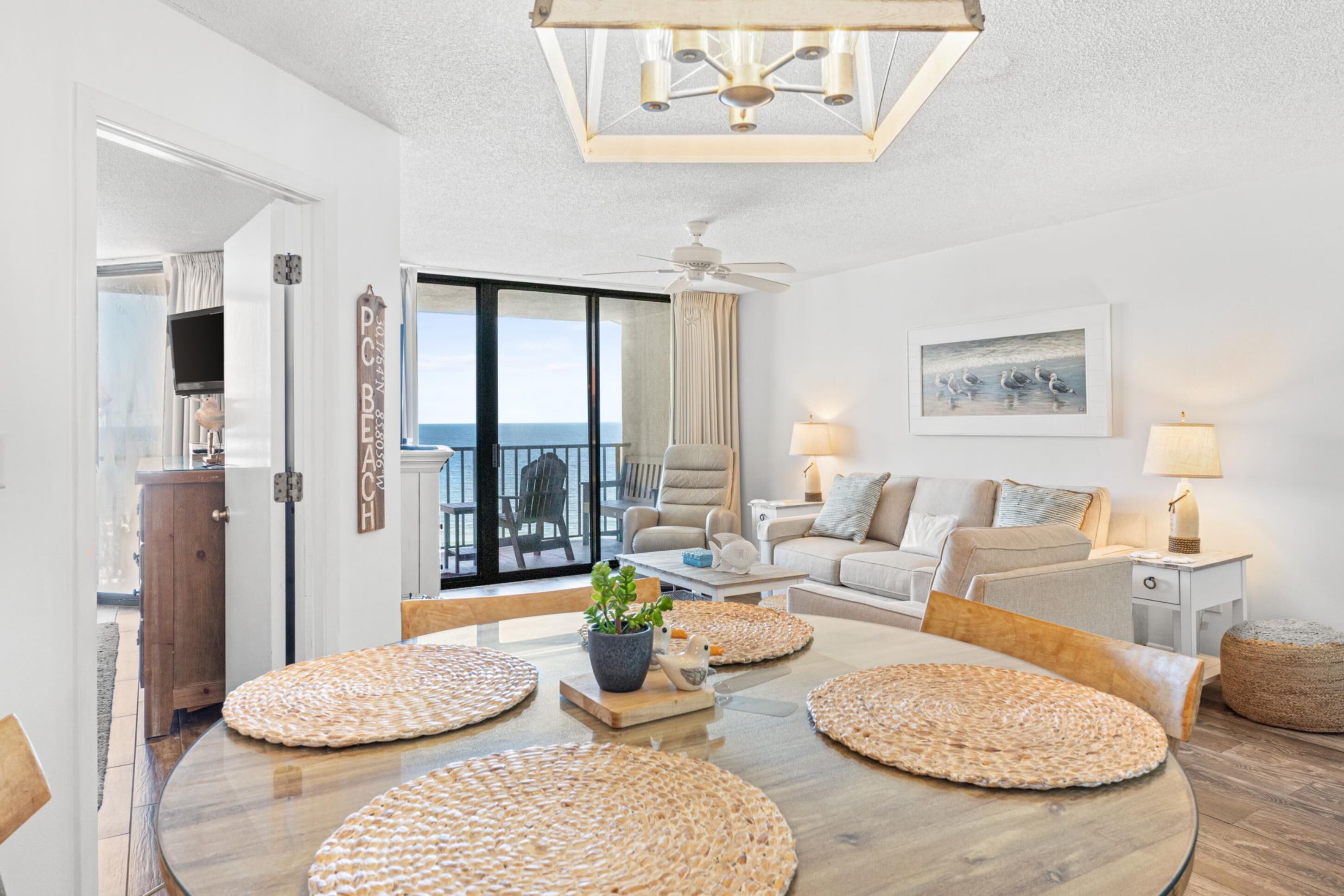 A modern living room with a round dining table, two armchairs, a sofa, a ceiling fan, and a large window with a view of the ocean.