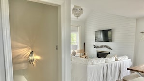 Une pièce avec un canapé blanc, un téléviseur à écran plat fixé au mur, un lustre et une cheminée.