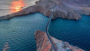 Uma estrada costeira serpenteando ao longo de uma linha de costa rochosa, com o pôr do sol ao fundo.
