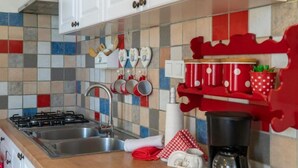 Uma cozinha com fogão a gás, pia e cafeteira. Há decorações em formato de coração na parede e uma prateleira vermelha com arabescos brancos.