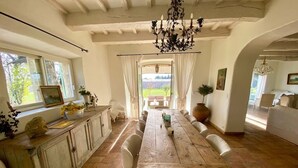 A dining area with a wooden table and chairs, a chandelier, and a view of the outdoors through large windows. - Mas Provençale Entouré de Pommiers, Piscine et Terrain de Tennis (Cavaillon)
