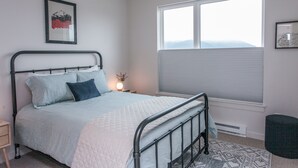 Une chambre à coucher avec un lit, une table de chevet, une fenêtre avec des stores et un tableau encadré au mur.