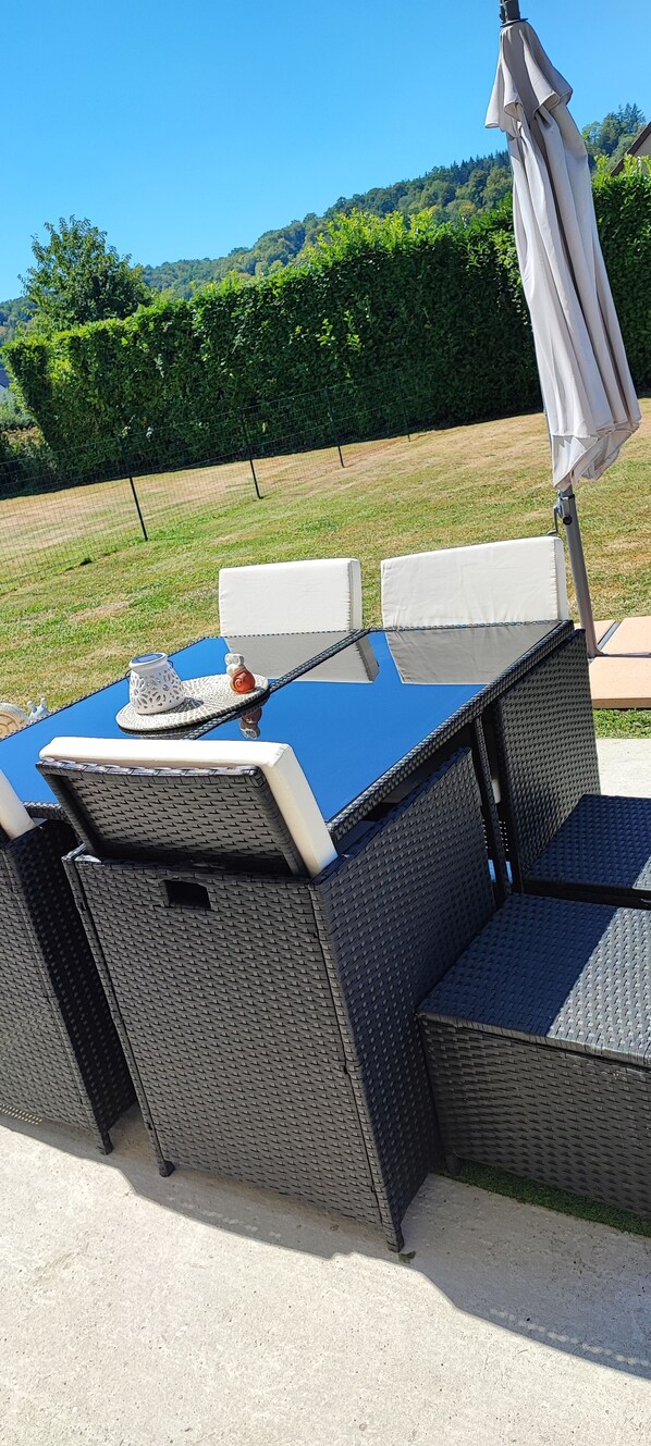 An outdoor dining area with a blue table and wicker chairs, an umbrella, and a view of a green landscape.
