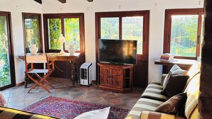 Ein gemütliches Wohnzimmer mit Holzboden, einem Fernseher, einer Couch, einem Esstisch mit Stühlen und großen Fenstern mit Blick auf viel Grün.