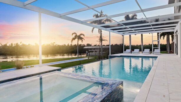 A modern outdoor pool area with clear views of a sunset over a body of water, surrounded by palm trees and a white pergola structure.