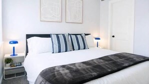 Un dormitorio con una cama, dos mesitas de noche con lámparas, una puerta y dos cuadros en la pared.