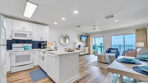 Eine moderne Küche mit weißen Schränken, einem eingebauten Backofen und einer Mikrowelle. Es gibt einen Essbereich mit Tisch und Stühlen, einen Balkon mit Meerblick und einen Deckenventilator.