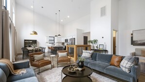 Un vaste salon avec un canapé gris, un fauteuil en cuir marron et une table basse. On aperçoit, à l’arrière-plan, une cuisine avec des meubles blancs et un coin repas comprenant une table et des chaises.