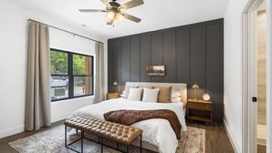 Une chambre avec un grand lit, un ventilateur de plafond, une table de chevet avec une lampe et une vue sur l’extérieur.
