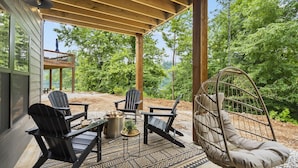 Un espace extérieur aménagé avec des sièges, abrité et doté de fauteuils Adirondack et d’un fauteuil suspendu, le tout entouré d’arbres.