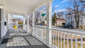 A covered porch with a white railing, a chair with a yellow cushion, and a flag with a green maple leaf and a star.