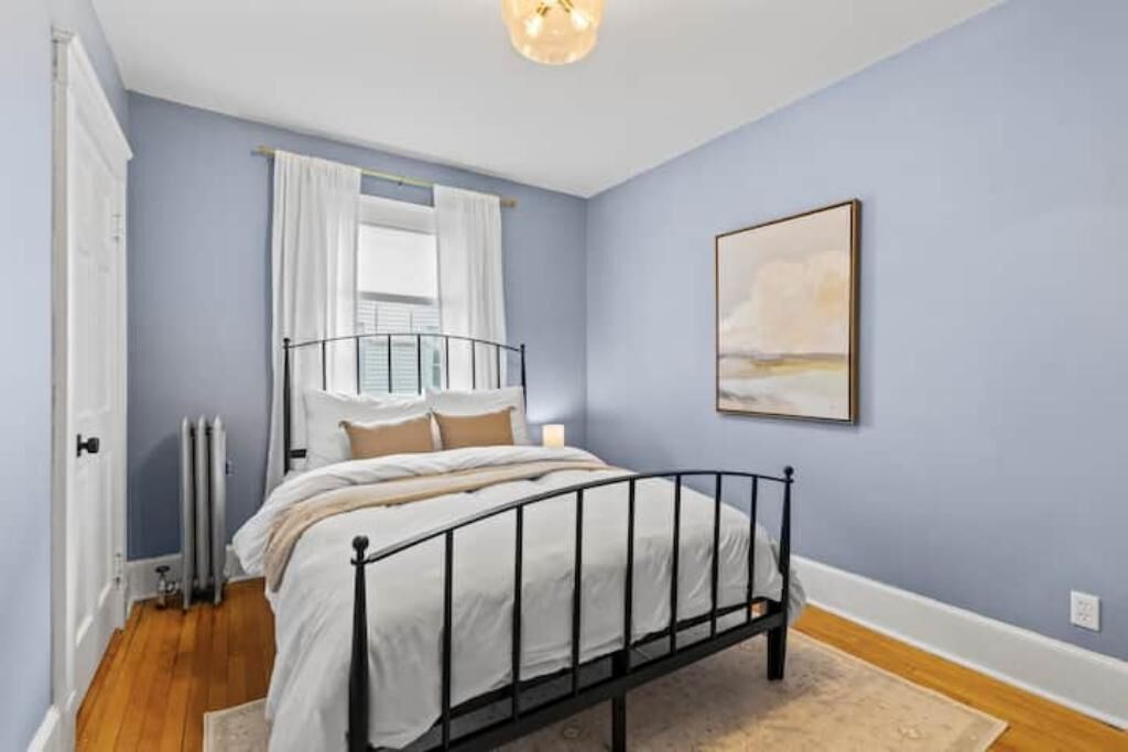 A bedroom with a bed, a window with curtains, a radiator, and a painting on the wall.