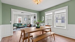 A dining area with a wooden table and benches, a chandelier, and a view of a green room through a window.